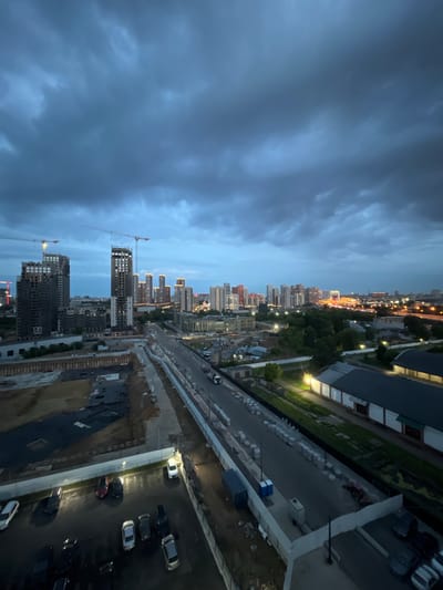 The image depicts a twilight cityscape with a cloudy sky. In the foreground, a construction site is visible with dark soil and scattered materials. Adjacent to the construction site is a paved area with several parked cars, some of which are wet, suggesting recent rain. Further into the scene, a road runs alongside buildings and a grassy area. A line of light-colored objects, possibly construction materials, is neatly arranged along the side of the road. In the background, a sprawling city skyline is illuminated by artificial lights, with numerous tall buildings of varying designs and heights. Construction cranes are visible among some of the buildings, indicating ongoing development. The overall atmosphere is subdued due to the low light conditions and cloudy sky. The location is identified as Chelyuskinsky, Russia.
