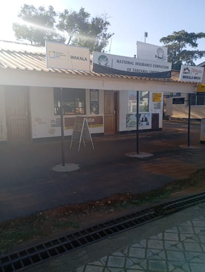 The image captures an outdoor commercial scene in Namanga, Kenya, bathed in bright sunlight, suggesting either morning or late afternoon due to a prominent lens flare in the upper left. No people are visible.
The scene features two adjoining single-story buildings with white-painted walls and corrugated metal roofs. The building on the left has a wooden door on its far side and an open service window area. The right-hand building also has a wooden entrance door. In front of the buildings, two dark poles stand on concrete bases, extending upwards, possibly for an awning or overhead structure.
Several signs are prominently displayed. On the roof of the left building, a sign reads "Tanzania Commercial Bank" next to the word "WAKALA" (meaning "agent" in Swahili). Above the right building, a large banner-style sign announces "NATIONAL INSURANCE CORPORATION OF TANZANIA LIMITED" with the "NIC" logo. To the far right, another sign displays "Equity," "Posta," and "WAKALA MKUU" (Main Agent). A smaller, A-frame sign in the foreground, near the left building, shows the word "vista." Additionally, a sign affixed to the right building clearly states "NO PARKING."
The ground in the foreground consists of a dark, paved surface, likely asphalt, with a narrow strip of dirt and sparse grass alongside it. A long metal drainage grate spans the bottom of the frame, separating the paved area from a patterned concrete section. Trees and other structures are visible in the background under the clear sky.