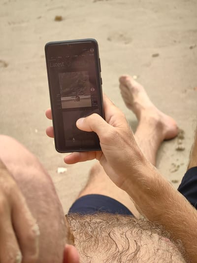 A man sits on a beach, viewed from an above angle, holding a dark-colored smartphone in his right hand. The phone screen displays what appears to be a social media or media-sharing application, featuring the words "Latest Created Inve" at the top and categories like "My," "New," "Top," "Core," "PPL," and "Near." An image is visible on the screen, with overlaid information indicating "2008 PATTH," "3 seen," "1 clicked," "0 saved" alongside details like "04/13/26 18:43" and the location "Hai Phong, Vietnam." The man's feet are visible on the sandy beach. The man is wearing dark blue shorts.