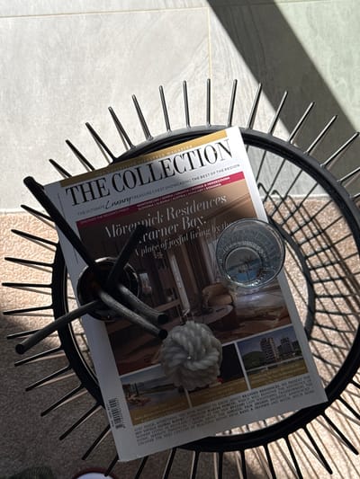 In the city of Bakovići, Montenegro, an open magazine titled "THE COLLECTION" lies on a circular black metal stand. The magazine, featuring "The Ultimate Luxury Treasure Chest Showcasing the Best of the Region," displays headlines like "Mövenpick Residences, Karner Bay: A place of joyful living by the sea" and "Introducing RIVIERA MONTENEGRO SWISS HOTEL: The New Mediterranean Dream." A glass with liquid and ice sits atop the magazine, reflecting the overhead light. Three dark, cylindrical objects, possibly incense sticks or decorative elements, are positioned on the stand and magazine. A small, white, textured decorative sphere is also present. The scene is illuminated by natural light, casting shadows across a tiled floor. The overall impression is one of a sophisticated and tranquil domestic setting.