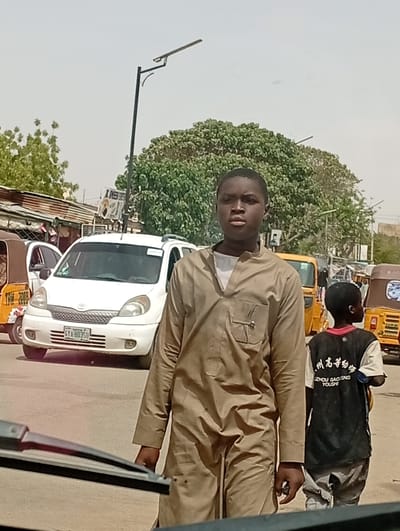 This image depicts a street scene in Hotoro North, Nigeria, under bright daylight conditions. In the central foreground, an adolescent male with dark skin and short hair is positioned, gazing forward. He is dressed in a light brown long-sleeved tunic, revealing a white undershirt collar, and holds a small dark object in his right hand. Behind and to the right of this primary subject, a younger male child walks away from the viewer, wearing a dark t-shirt with white text, including "ZHOU GAOENG YOUSHI" and Chinese characters, and light-colored trousers.
A white Toyota Sienta minivan, bearing Nigerian license plate GTA-803SU, is situated in the midground behind the primary subject. Multiple yellow commercial tricycles are present on the paved road; one is partially visible on the far left, another further in the background, and a third on the far right. Low-rise structures with corrugated metal roofs are visible on the left background, alongside several green trees, including a large, dense specimen. A streetlight with an extended arm and a solar panel is present in the upper frame against a pale sky. A partial windshield wiper in the lower left corner indicates the image was captured from inside a vehicle.