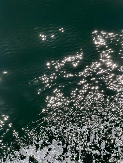 The image depicts a close-up, top-down view of dark green water, likely the ocean, with a section of foamy white water in the foreground. Sunlight reflects off the surface of the water, creating numerous bright, star-like sparkles across the scene. The water appears slightly rippled, suggesting gentle movement. The image does not contain any people, identifiable structures, or specific landmarks that would indicate its precise location within Point Roberts, United States, beyond it being an outdoor aquatic environment.