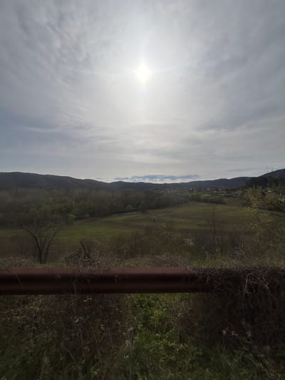 The image shows a landscape view in an outdoor setting. A bright sun is visible in the cloudy sky on the left side. A mountainous region extends from the left towards the center. A field area with green and brown vegetation is observed to the right of the mountains. There's a dark vertical pole-like structure on the right, adjacent to more dense vegetation. The specific location in Kosti, Bulgaria, is not identifiable from the image. No people are visible.