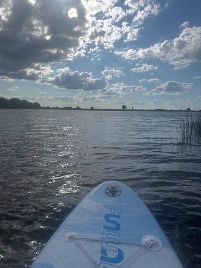 The image shows a close-up view of the front of a blue and white stand-up paddleboard on a body of water. The paddleboard has a logo that reads "FUNWATER" in blue, capitalized letters, and some white circular fixtures. The water is dark with ripples and reflections of the sky. In the background, a line of trees and reeds can be seen along the distant shore. The sky is a mix of blue with white and gray clouds, suggesting partly cloudy weather. The sun is shining, creating bright highlights on the water. The perspective is from someone on the paddleboard looking forward and slightly to the left. The overall mood is peaceful and serene, indicative of a recreational activity on a pleasant day. The scene appears to be in Russia, based on the provided location context.