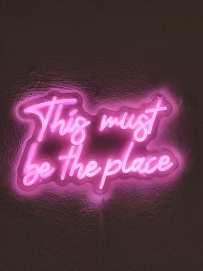 A bright pink neon sign reading "This must be the place" is mounted on a textured, dark wall. A power cord extends down from the sign. The background appears to be a dark-colored wall with a rough texture. The location details are not evident from the image.