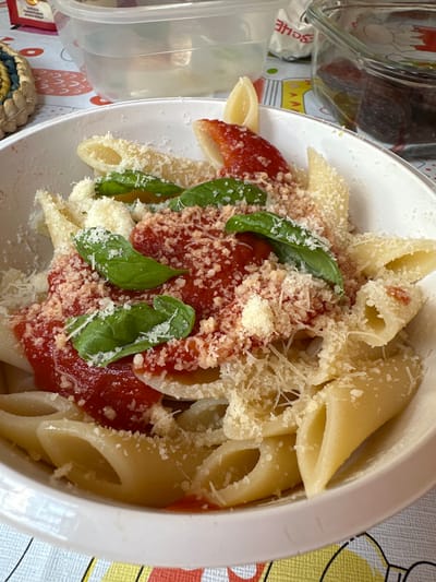 A white bowl filled with penne pasta, topped with marinara sauce, shredded Parmesan cheese, and fresh basil leaves, is centered on a table. The table is covered with a colorful, patterned tablecloth. In the background, out of focus, are several containers, including a clear plastic container with water and a glass container with dark food items. The image does not provide any information about the city of Borno, Italy, or its location.