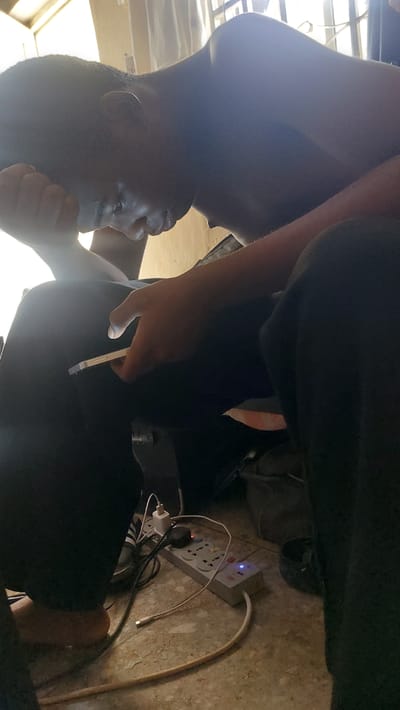 In New Nyanya, Nigeria, a person, partially visible and hunched over, is seen from the side and slightly from above. They are wearing dark clothing and have their hand resting on their head, appearing to be in a pensive or stressed posture. A smartphone is held in their other hand, with the screen illuminated. The background reveals a dimly lit interior with a window covered by curtains. On the floor, a power strip with several plugs and cables is visible, connected to electrical devices. The overall impression is a candid moment of someone engaged with their phone in a modest indoor setting.