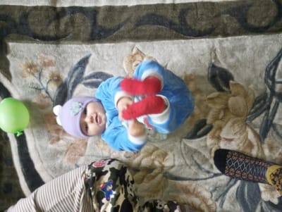 An overhead view depicts an infant lying on its back on a patterned carpet or rug within an indoor environment in Galaosiyo, Uzbekistan. The carpet features a beige and light brown base with dark brown/black and lighter floral or abstract designs, along with striped sections on the left and top edges.
The infant is dressed in a light blue fuzzy romper with white trim, bright red socks, and a light purple knitted beanie adorned with a pale green decorative element on the forehead. Its mouth is slightly open, and it is looking directly upwards towards the camera, with arms bent and hands raised towards its chest, and legs bent with feet elevated.
A deflated or partially deflated bright green balloon rests on the carpet to the left of the infant's head.
Parts of an adult's lower body are visible in the foreground. The left leg wears light beige and white striped pants. The right leg wears camouflage-patterned pants featuring a blue airplane patch on the knee area, and the right foot is clad in a black sock with a diamond and floral geometric pattern in white, light blue, and orange/brown.