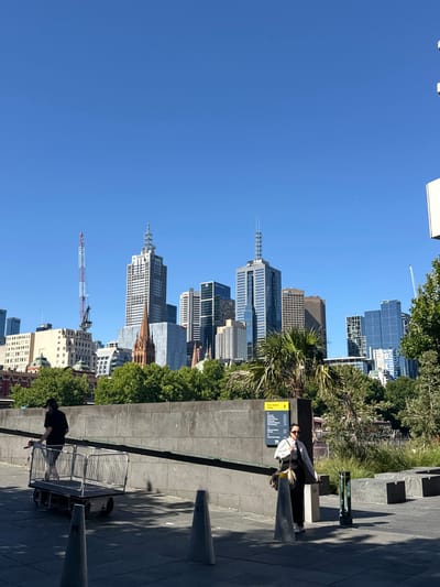 A cityscape dominates the left, with buildings rising against a clear blue sky. Lush green trees intermingle with the urban landscape. A concrete structure occupies the right, featuring cones and a person sitting on a bench. Below, another person pushes a metal cart along the walkway. The scene is bathed in bright sunlight.