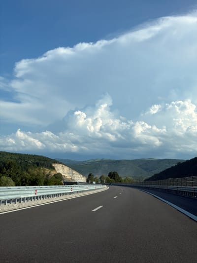 The image depicts a highway scene in Peshterna, Bulgaria, under a dramatic sky filled with large, white, fluffy clouds. The highway itself is paved and dark, with a white dashed line marking the lane. On the left side of the road, a metal guardrail with red reflectors runs along the edge. Beyond the guardrail, a quarry is visible, with layered rock formations and some trees growing on the slopes. The background features rolling hills covered in lush green vegetation, with some darker, more distant mountains. The overall impression is one of a vast, open landscape with a sense of natural beauty and development.