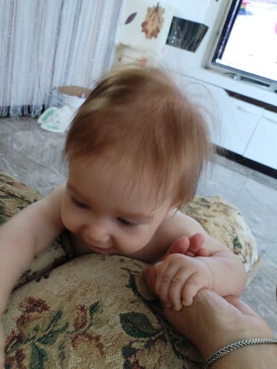 A close-up, over-the-shoulder view shows a baby lying on a patterned fabric surface, possibly a sofa cushion. The baby is facing downwards, with its head turned to the right, and its eyes are closed. The baby's hair is light brown and somewhat messy. One of the baby's hands is resting on the fabric, and another hand, presumably belonging to an adult, is holding the baby's fingers. The adult's hand is adorned with a silver bracelet. The background is out of focus but suggests an indoor setting with furniture and a television screen visible. The lighting appears to be natural, coming from the left side of the frame. No specific details about the city of Vinogradny, Russia, are discernible from the image itself.