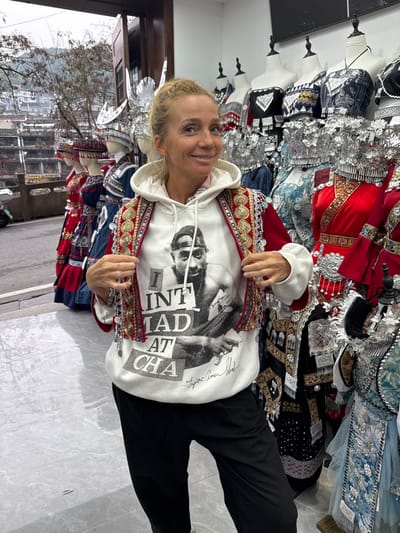 An adult female, fair-skinned with light brown hair pulled back, stands smiling directly at the camera, holding the front edges of a red, heavily embroidered traditional-style vest adorned with gold-colored embellishments and silver coin-like pendants. She wears a white hooded sweatshirt featuring a black-and-white image of Tupac Shakur and the text "I INT MAD AT CHA" along with his name, and plain black trousers.

The scene takes place inside a shop in Fenghuang, China, with a large window in the background looking out onto the city. The exterior view reveals traditional stilt houses and buildings typical of Fenghuang, with a misty hillside and trees under an overcast sky. Inside the shop, behind the woman, numerous mannequins are visible, displaying elaborate traditional Miao/Hmong clothing, including vibrant red, blue, and black garments with extensive silver ornamentation, embroidery, and ornate silver headpieces. The shop floor is light gray tile, and the interior walls are white.