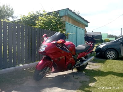 A red Honda CBR1100XX Super Blackbird motorcycle is parked on a gravel and grass surface next to a grey wooden fence. The motorcycle is facing left and is equipped with a black windscreen and a black top case. Behind the fence is a light-colored wooden structure with a blue door and trim, partially covered by green vines. To the right of the motorcycle, a dark-colored car is visible, parked on the grass. The sky is partly cloudy, and the scene appears to be in a rural or suburban setting. A date stamp at the bottom right corner reads "21.06.2026 14:26".