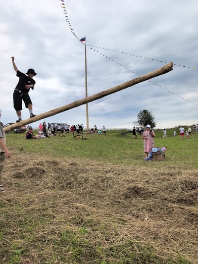 The image depicts an outdoor event, likely a festival or competition, taking place in a grassy field in Koyanovo, Russia, under a cloudy sky. A long, thick wooden log is positioned diagonally across the frame, with one end resting on the ground and the other elevated, supported by a tall wooden pole. A young man wearing a black t-shirt, black shorts, white socks, and a black hat is balancing precariously on the log, with his left arm raised. He is facing towards the left of the frame.
In the background, a crowd of people is gathered, some sitting on the grass and others standing. A tall pole with a Russian flag is visible in the distance. Colorful triangular flags are strung between poles and appear to be decorations for the event. Several vehicles, including a white van and a red car, are parked in the background.
To the right of the frame, an older woman wearing a pink dress and a white hat stands near a cardboard box filled with items, possibly prizes or supplies. She is holding a smartphone in her hand. Further in the background on the right, other people, including children, are visible mingling. The foreground of the image is covered with dry hay and green grass, suggesting a rural or semi-rural setting. A bicycle lies on its side in the middle ground.