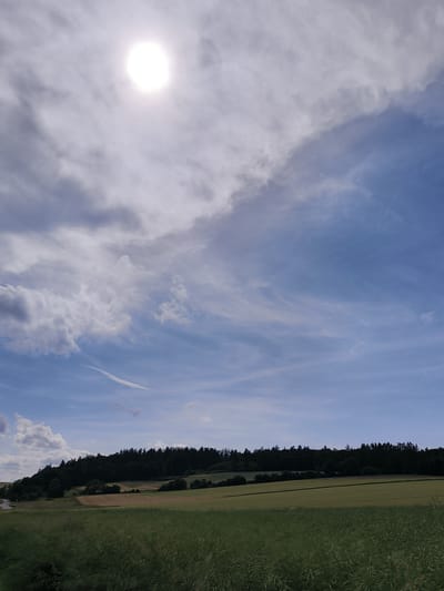 The media file depicts a panoramic view of a rural landscape under a dynamic sky. The sun, a bright white disc, is partially obscured by wispy clouds in the upper left quadrant, suggesting it is likely midday or late afternoon. The sky transitions from bright, scattered clouds near the sun to a clearer, pale blue with streaks of cirrus clouds stretching across the frame. A thin contrail from an aircraft is visible as a faint white line in the upper part of the image.
The foreground is dominated by a field of tall, green grass, appearing slightly blurred, suggesting a gentle breeze. Behind this, there are rolling hills with patches of farmland, including areas of golden stubble or dry crops, and vibrant green fields. The mid-ground is characterized by a densely wooded hillside, forming a dark, undulating treeline against the lighter sky. To the far left, a winding road with a single vehicle can be seen on the edge of the frame, hinting at the presence of human activity in the vicinity.
The overall mood is serene and natural, with the bright sun and vast sky conveying a sense of openness. The location is identified as Sebbeterode, Germany. There is no visible text in the image.