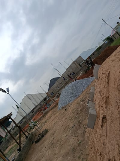This image depicts a construction site in Masaka, Nigeria, under a cloudy sky. The foreground is dominated by a large pile of fine, light brown sand and a few concrete blocks. Behind the sand, a substantial mound of light gray gravel is visible. In the mid-ground, a multi-story building is under construction, with visible brickwork and scaffolding. Several workers are engaged in activity; one is pushing a wheelbarrow, another appears to be working near the wheelbarrow, and others are visible on different levels of the building. A streetlamp stands to the left of the frame, illuminating the scene. Further in the background, more buildings are present, some with corrugated metal roofs, and a utility pole with overhead wires is visible. The ground is a mix of dirt and some sparse vegetation. The image is tilted, suggesting it was taken at an angle.