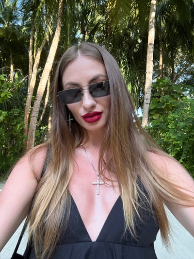 The image shows a close-up, portrait-style selfie of a woman with long, blonde hair. She is wearing sunglasses and has red lipstick. A silver necklace with a cross pendant is visible around her neck. She is wearing a black top, and her skin appears slightly shiny, suggesting she might be in a warm, possibly humid environment.

The background consists of lush green foliage and palm trees, with some dappled sunlight filtering through. This suggests an outdoor setting, likely in a tropical or subtropical location. The presence of palm trees and the mention of "Maldives" as a location cue strongly point towards a beach or island setting. The lighting and the bright green of the leaves suggest it is daytime, possibly late morning or afternoon.

The woman's pose and direct gaze into the camera are typical of a selfie. Her facial expression, with her lips slightly parted and makeup applied, could be interpreted as playful or confident. There is no visible text in the image. The overall impression is one of a casual, vacation-like moment captured outdoors in a tropical environment.