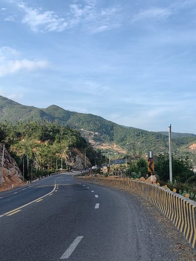 The image displays a mountainous road segment located in Tuy Phong 4, Vietnam, under a clear blue sky with scattered cirrus clouds. The asphalt road, dark grey with white dashed and double yellow center lines, curves sharply to the right in the foreground and then gently to the left into the distance. A yellow and black striped concrete barrier, topped with small yellow reflectors, lines the outer edge of the right-hand curve. A yellow and black chevron sign, indicating a sharp left turn, is visible on the right, positioned behind the barrier, adjacent to a black and white striped warning sign. The left side of the road is bordered by a steep, rocky embankment composed of reddish-brown rock and covered with various green trees and shrubs. Multiple utility poles with wires run alongside the road on both sides. In the mid-ground, a white sedan and two motorcycles are observed traveling on the road, moving away from the viewer. The background features several layers of prominent mountains and hills, densely covered in green forestation, with some areas exhibiting lighter or sparser vegetation.