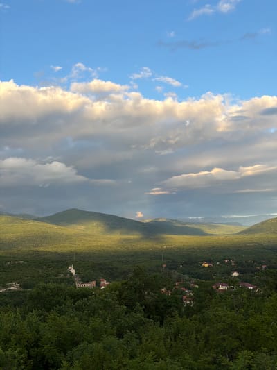 The image displays a vast, serene landscape under a dynamic sky, likely captured during late afternoon or early evening. The upper left portion of the frame features a clear, bright blue sky, which transitions towards the right into an impressive expanse of voluminous white and grey clouds. The edges of these clouds are brilliantly illuminated by the low-lying sun, creating a sense of depth and ethereal beauty.

Below the sky and clouds, the terrain consists of rolling hills and mountains, all densely covered in lush green vegetation, primarily trees. The sunlight beautifully bathes sections of these slopes, creating vibrant highlights and long, soft shadows that accentuate the natural contours of the land. In the mid-ground and extending towards the right side of the image, a dense forest fills the foreground.

Scattered within these wooded hills, particularly on the right side of the mid-ground, are several small buildings, appearing to be houses or a small community. These structures are nestled amongst the trees, with visible roofs often in shades of red or lighter colors, blending into the natural environment. There are no people visible, nor are there any discernible activities taking place. The scene exudes a tranquil and majestic atmosphere, characteristic of a mountainous region. The warm, soft lighting underscores the peaceful transition of the day. No visible text is present in the image. The location is Vidrovan, Montenegro.