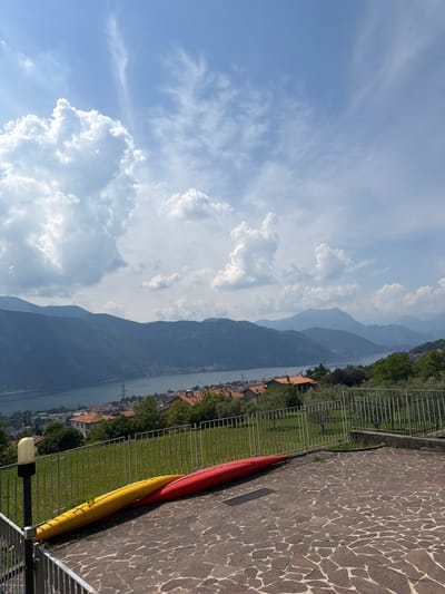 A high-angle, medium shot captures a serene landscape in Abbadia Lariana, Italy, on a bright, partly cloudy day. The foreground is dominated by a paved stone patio where two kayaks, one bright yellow and the other red, lie on their sides, angled towards the left. A metal fence runs horizontally across the middle of the frame, separating the patio from a grassy slope beyond.
The mid-ground reveals a picturesque town nestled along the edge of a large, calm body of water, likely Lake Como, given the location. Red-tiled rooftops are visible amongst lush green trees. In the distance, a series of imposing, hazy mountains form a majestic backdrop, their peaks fading into the sky. The sky itself is a vibrant blue, adorned with scattered white cumulus clouds, some of which are large and billowy, catching the sunlight. A single utility pole with wires is visible on the slope leading down to the lake. The overall atmosphere is peaceful and idyllic.