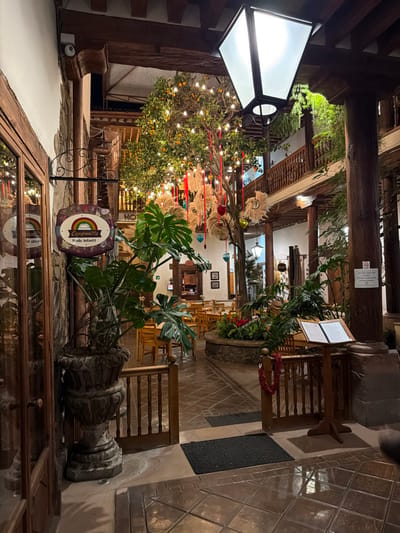 An ornately decorated interior space is visible. A white-lit, boxy lamppost is mounted on a wood beam. A leafy, lighted tree stands near a small sign bearing the words "compania infantil". Various decorations are suspended from the ceiling. Tables and chairs populate the background, while stone-tiled floors lead further into the space. Lush plants in large, decorative pots add to the ambiance.