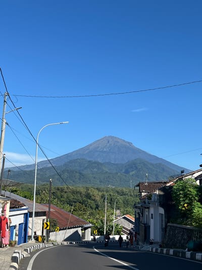 The image presents a scenic view of a prominent mountain, likely a volcano, under a clear, bright blue sky. The mountain dominates the left side of the frame, with its slopes covered in dense green vegetation that becomes hazier towards the peak, suggesting distance. A faint, thin cloud wisp is visible against the otherwise cloudless sky.
In the foreground and along the right side, a paved road curves gently, delineated by a black and white striped curb. Several buildings with varied roof materials, including corrugated metal and brown tiles, are situated along the roadside, surrounded by abundant green trees and bushes. Utility poles with overhead power lines crisscross the lower part of the image, and a modern white streetlamp curves upwards into the sky.
Notable details include several yellow and black chevron road signs indicating a curve to the left. On the bottom right, colorful laundry is hanging to dry, with part of a motorbike also visible nearby. Two satellite dishes are mounted on rooftops. The scene is bathed in bright sunlight, indicating it is daytime, likely mid-morning or early afternoon. The overall impression is one of a peaceful, everyday setting in a community nestled close to a significant natural landmark, consistent with the location context of Magelang, Indonesia. No explicit text is visible beyond the pictorial road signs.