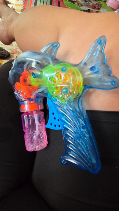 A transparent blue, fish-shaped bubble gun with orange and green internal components rests on a person's lap, who is wearing black clothing. A pink bottle filled with soapy water is attached to the gun. The background includes a colorful, striped fabric. The image does not provide any information about the city of Vozsiiatske, Ukraine.