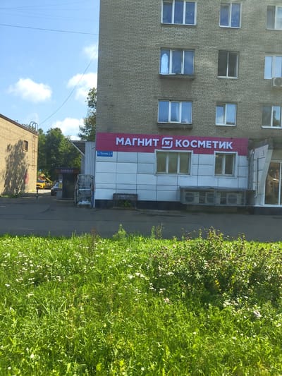 The image shows the storefront of a "Magnit Kosmetik" store located in the city of Zhukovsky, Russia. The store is situated on the ground floor of a multi-story brick building. The storefront features a large, prominent sign with the store's name in white lettering on a pink background. Below the main sign, there is a smaller blue sign that reads "ул. Чкалова, д. 6", indicating the street address is Chkalova Street, building number 6. The building itself has multiple windows on its upper floors. In front of the store, there are several air conditioning units installed under a sheltered area. To the left of the store, there is another building, and a yellow car is visible further down the street. The area in front of the buildings appears to be a paved walkway or street, with a grassy area in the foreground. The sky is blue with some clouds, suggesting it is daytime.