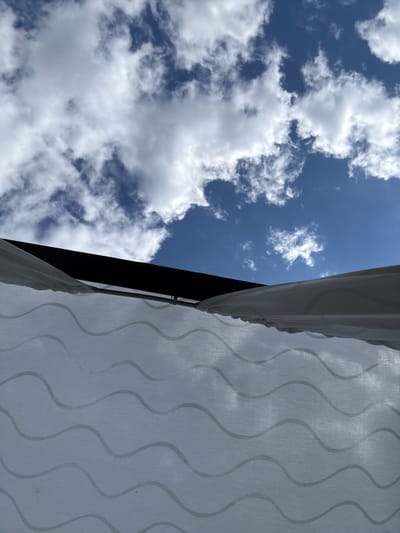 The image depicts a low-angle view looking up towards the sky. The foreground is dominated by a white fabric with a subtle wave pattern, possibly a curtain or canopy. A dark, horizontal structure, likely the edge of a roof or awning, is visible above the fabric. The background consists of a bright blue sky with scattered white clouds. The scene does not provide any specific visual cues to indicate its location in Nechkino, Russia. There are no people, actions, or interactions visible in the frame.