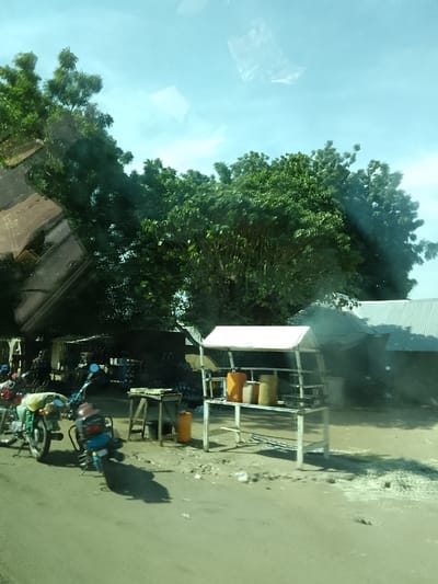 The image depicts a roadside scene in Minting Chip, Nigeria, under daylight conditions. The foreground features a dark, likely paved, surface. On the left, two motorcycles are parked; one is a dark color with substantial luggage tied to its rear, and the other is blue. Further left in the background, several indistinct figures are visible amidst what appears to be a gathering or market activity.

In the mid-ground on the right, a white-framed stall with a white peaked roof stands prominently, displaying several jerrycans of varying colors (orange and yellow) on its shelves. To the left of this stall, a wooden table with items on its surface functions as another makeshift vendor stand. Directly behind the white stall and to its left, multiple simple structures with corrugated metal roofs are visible. Smoke is actively rising from an area behind these structures, suggesting a source of combustion. Dense green foliage from large trees dominates the upper and mid-sections of the background, extending across the scene and partially obscuring the distant sky, which appears light blue. Reflections are visible across the upper and central portions of the image, indicating it was captured through a window or glass.