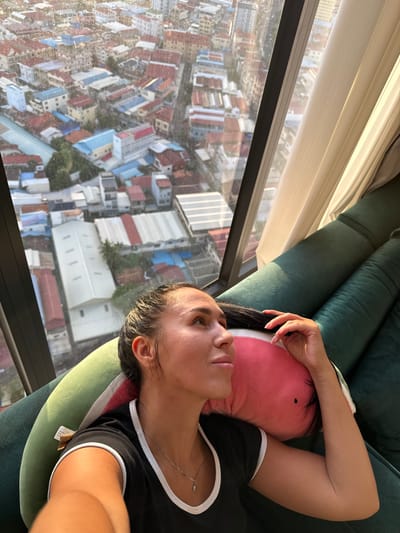 The image shows a woman lying on a dark green couch, resting her head on a pink and green pillow. She is looking upwards and to the right, with her right hand touching her hair. The woman is wearing a black t-shirt with white trim and a silver necklace. Behind her, a large window overlooks a densely populated urban area with buildings of various colors and sizes, including residential homes and industrial-looking structures. The view appears to be from a high vantage point, suggesting the location is an elevated apartment or building within the city of Khan Mean Chey, Cambodia. The lighting suggests it is daytime, with sunlight casting shadows on the couch and the woman.