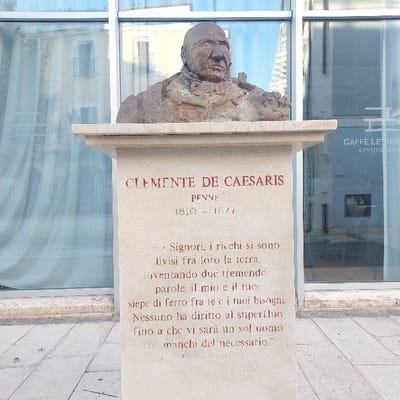 A weathered bronze bust of Clemente De Caesaris, depicting an older man with a balding head and formal attire, stands prominently on a light-colored stone pedestal. The pedestal's front face is inscribed in red capital letters with "CLEMENTE DE CAESARIS," followed by "PENNE" and the life dates "1810 - 1877." Below the dates, a quote in Italian is carved, translating to: "For the Lords, the rich have divided the earth among themselves, inventing two tremendous words, mine and yours: an iron fence between you and your needs. No one has the right to superfluity as long as there is even one man who lacks the necessary." This is followed by "Pastor di Popolo." The statue is situated outdoors on a paved plaza with light-colored square tiles. Behind the statue is a modern building featuring large, reflective glass windows. The windows reflect an older building with balconies and the sign for "CAFFÈ LETTERARIO CINQUE SENS." The scene is brightly lit by natural light. This monument is located in Piazza Emilio Alessandrini, Pescara, Italy.