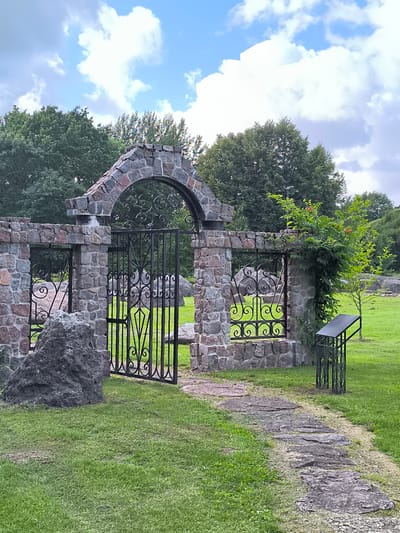 This image depicts an ornate stone gate with wrought iron inserts, set within a park-like environment. The gate is constructed from rough-hewn stones of varying sizes and colors, including shades of gray and pink, forming an arched opening. A large, dark boulder sits to the left of the gate. To the right, a path made of flat stones leads away from the gate through a manicured lawn. Beyond the gate, more large boulders are scattered across the grassy expanse, and a small tree with red berries is visible. The background is filled with lush green trees, suggesting a temperate climate. The sky above is blue with fluffy white clouds, indicating fair weather, likely during the daytime. There is a dark metal signpost to the right of the gate, but no visible text is discernible. The overall scene conveys a sense of rustic beauty and tranquility.