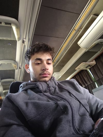 The image displays a young adult male seated within the interior of a commercial passenger vehicle, located in Mankat al-Hatab, Syria. The male, centered in the frame, has short, dark, curly hair and light facial hair. He wears a dark grey zippered hooded sweatshirt with visible drawstrings at the collar. A white wireless earbud is present in his right ear. His head is slightly tilted forward, and his eyes are cast downwards. The vehicle's overhead area features alternating dark grey textured panels and light grey structural elements. A linear LED light strip illuminates the ceiling, and a rectangular air vent is visible overhead on the left. On the right, brown pleated curtains are drawn, indicating a window, and a light-colored overhead storage compartment is partially visible. The male occupies a dark-colored seat.