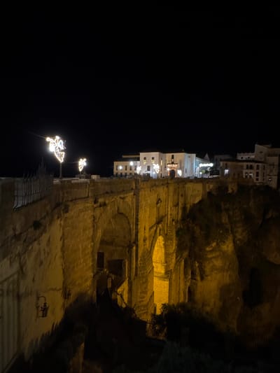 A nighttime view of a cliffside village with buildings illuminated by string lights and decorative figures. The buildings are primarily white, contrasting with the dark sky. The cliff face is textured and layered, lit by warm yellow lights that highlight its form. A small figure, possibly a sculpture, stands in the lower right corner. Overall, the image captures a serene and unique atmosphere.