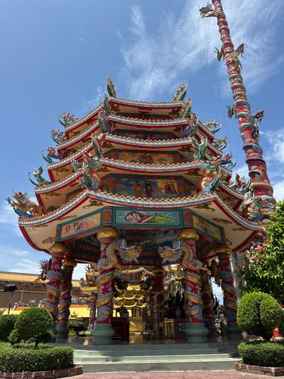 A multi-tiered, ornate temple structure with dragon motifs and brightly colored decorations stands under a clear blue sky with scattered clouds. A tall, red, dragon-entwined pillar is visible to the right of the temple. The temple is surrounded by lush green bushes and trees, with paved walkways and steps leading up to it. The background shows more temple structures with orange roofs. The scene is located in Ang Sila, Thailand. There are no people visible in the image.