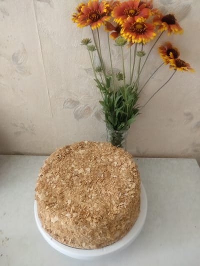 A round cake, covered in crumbled pastry, sits on a white cake stand. The cake stand is on a light-colored countertop, which is in front of a light-colored, textured wall. To the right of the cake, a bouquet of bright orange and yellow flowers with dark centers is displayed in a clear glass vase. The flowers are arranged with green stems and some unopened buds. The overall scene appears to be indoors, possibly on a table in a room. The location is not specified, and there is no information about the city of Ananiv, Ukraine, or any activities taking place.