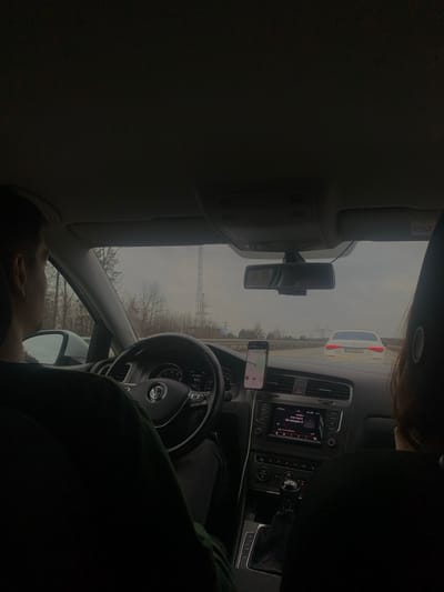 The image captures the interior perspective from the rear seat of a Volkswagen vehicle in Oliivka, Ukraine, looking forward through the windshield. A male driver, visible from the left side of his head and shoulder, wearing a dark top, grips the Volkswagen steering wheel. His left hand is positioned on the wheel. A front passenger, with dark hair, is partially visible from behind on the right side of the frame. A smartphone displaying a navigation application is mounted on the dashboard to the right of the steering wheel. The vehicle's infotainment system in the center console is active, showing a media interface. Beyond the windshield, a multi-lane road is visible under an overcast sky. A white sedan, with its brake lights illuminated, is directly ahead in the right lane. Bare-branched trees line the roadside, and a tall, lattice-style electrical or communication tower stands prominently in the distance to the left of the road. A smaller pylon-like structure is also visible further down the road on the right.