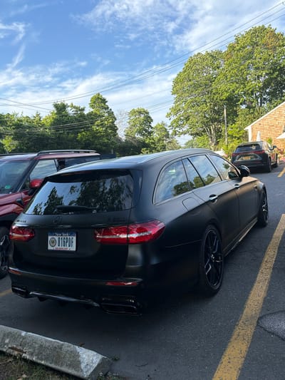 In Bridgehampton, United States of America, a matte black Mercedes-Benz E63 S Wagon is parked in a lot. The car features a New York license plate that reads "816PDF". It is parked to the right of a red SUV. In the background, a dark-colored SUV is parked further down the lot, and a building with a shingled roof is visible. Trees and overhead power lines are present in the scene. The ground is asphalt, marked with yellow lines. The sky is blue with scattered white clouds.