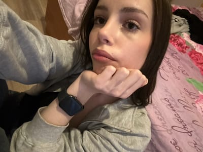 A young woman with dark hair is depicted in a close-up, angled view, lying down indoors. Her right hand, with fingers curled, supports her chin, partially obscuring the lower-right portion of her face. She has dark eyebrows and visible eyelashes. Her lips are slightly parted. She is wearing a light grey zippered hoodie over a dark garment. A black smartwatch with a rectangular face is visible on her left wrist. To her right, a light purple or pink patterned bedsheet or duvet cover features floral motifs and cursive text, with crumpled light and dark fabrics visible on it. To her left, a light brown surface is visible in the background. The scene is located in Волховец, Russia.