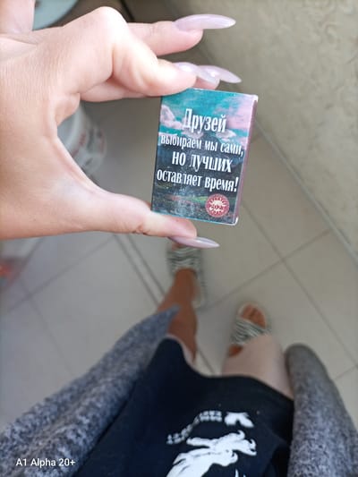 A first-person perspective shot shows a hand with long, manicured nails holding a small, rectangular box. The box has text in Russian that reads "Друзей выбираем мы сами, но лучших оставляет время!" which translates to "We choose friends ourselves, but time leaves the best!". Below this, there is a circular stamp with the year "2022" prominently displayed, along with other Cyrillic text. The background is out of focus but appears to be an indoor setting with tiled flooring and what looks like furniture or a doorway. The person's feet, wearing striped sandals, are visible in the lower part of the frame, suggesting they are standing. The overall scene is casual, possibly domestic. The location of Улукаўскі сельскі Савет, Belarus is irrelevant to the visual content of the image.