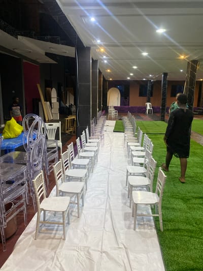 The image shows a large hall in a building located at the coordinates 4.037, 9.204 in the city of Limbe, Cameroon. The hall is being set up for an event. There are two rows of white chairs arranged in a long aisle, with white plastic sheets covering the floor between the rows. One woman is standing near the right edge of the image, looking down at the chairs. A man is walking away from the camera, out of focus, in the background of the image. There are many bright white lights illuminating the hall from the ceiling. The walls of the hall are black, white, and brown. There is artificial grass on the floor to the right of the white plastic sheets, and some clear plastic chairs are stacked in the left foreground. The image captures the preparation process for an event in the building, suggesting a celebration or ceremony is planned.