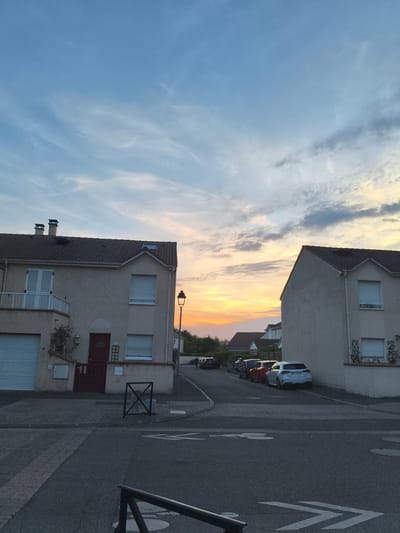 The image shows a residential street in Carrières-sous-Poissy, France, at dusk. Two houses are visible, one on the left and one on the right. The house on the left has a beige facade, a garage door, a red front door, and shutters on its windows. The house on the right also has a beige facade and shutters. Several cars are parked along the street. The sky is a gradient of blue and orange, indicating sunset. A lamppost stands on the sidewalk between the two houses. The street is paved with asphalt and has painted lane markings.