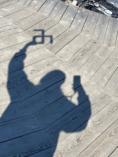 A shadow of a person holding a camera and taking a picture is cast on a wooden deck in Gubakha, Russia. The deck planks are laid in a herringbone pattern and are weathered gray. A small patch of snow is visible on the left edge of the deck, near some bare branches. The person's head is silhouetted with the camera held in their hand.
