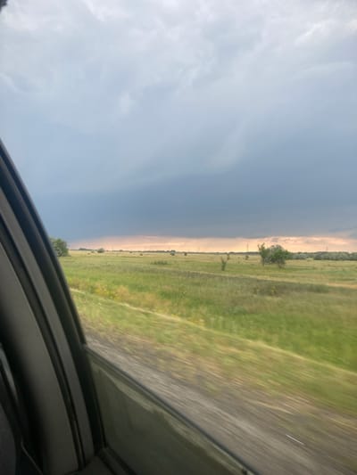 The image depicts a wide, open landscape with a dramatic contrast in the sky. On the left side, the sky is a deep, ominous gray, suggesting an impending storm or heavy cloud cover. This dark expanse sharply divides the scene from the right side, where the sky is lighter, appearing a hazy pale blue or yellow, with some wispy white clouds.
The ground below is a mixture of green fields and a road. The fields are lush and appear to be covered in grass, with some scattered yellow wildflowers visible. A road, likely paved, runs diagonally through the right side of the frame, showing motion blur, indicating it's being viewed from a moving vehicle. The perspective suggests the photograph was taken from inside a car, with the bottom of the frame showing part of the car's interior, possibly a window frame or sunroof.
In the distance, along the horizon where the dark and light skies meet, a line of trees is visible. There are also some scattered trees and utility poles dotting the landscape. The overall impression is of a vast, rural setting in Russia, perhaps near Rubtsovsk, experiencing changing weather conditions. The contrast between the dark, stormy sky and the brighter, hazy sky could suggest the edge of a weather front or a localized storm. The time of day is difficult to ascertain precisely due to the lighting, but the hazy quality of the lighter sky might suggest late afternoon or early evening. No people or specific objects are discernible.