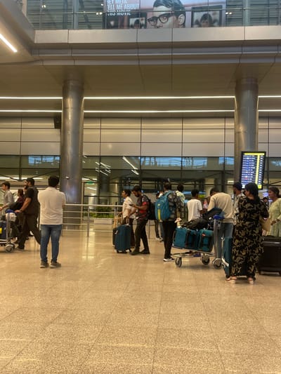 The image depicts an interior scene at Rajiv Gandhi International Airport in Hyderabad, India. Several individuals are visible, mostly standing and holding luggage or pushing trolleys. The environment is spacious and modern, with polished floors, large pillars, and overhead lighting. A digital display board showing flight information is positioned on the right side of the image. People are dressed in casual attire, and some are interacting or waiting. The overall atmosphere suggests a busy but orderly travel hub.