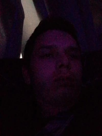 The image is a low-light, close-up selfie of a person's face, predominantly illuminated by purple light. The background is dark and indistinct, with some hints of lighter tones and textures. The person's mouth is slightly pursed, and their eyes are visible but somewhat obscured by the lighting. The overall impression is that of a selfie taken in a dimly lit environment. The location in the city of Павловка, Russia cannot be determined from the image.