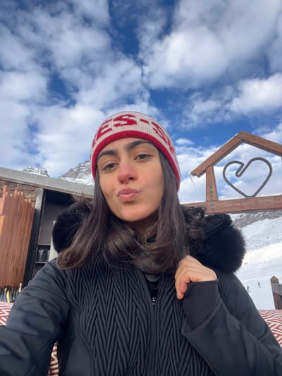 A female subject is depicted in a close-up, eye-level selfie against a partially cloudy blue sky. She possesses dark hair, worn down over her shoulders, and is wearing a red and white knitted beanie with visible text and a black winter jacket featuring a textured, chevron-like pattern on the torso and a black fur-trimmed hood. Her left hand is visible, gripping the cuff of her right sleeve. Her lips are pursed in a pouting expression.
The background reveals a snow-covered mountainous environment. To the left, a modern structure with vertical wooden or copper-toned slats is partially visible, with dark objects below it. To the right, a large wooden frame, shaped like a house outline, contains a metal heart emblem suspended within it, indicative of a ski resort landmark. Distant figures, likely skiers or snowboarders, are discernible on the snow-covered slope. The subject appears to be seated on a red and white patterned surface. The scene is located in a ski resort area in Valtournenche, Italy.