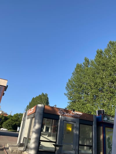 The image shows a clear blue sky over a street scene in Kiselyovsk, Russia. In the foreground, there are two kiosks. The kiosk on the left has a sign with Russian text that appears to say "Горячие" (Hot) and "Беляши" (Belyashi), which are traditional Russian pastries. The kiosk on the right has a sign with Russian text that says "доб" (short for "добро пожаловать" - welcome) and "Сладк" (short for "сладкое" - sweets). There are trees with green foliage visible behind the kiosks. A white car is parked on the street to the left of the kiosks, and a building with a beige facade is partially visible in the far left background. The scene appears to be set during daytime, with sunlight casting shadows.