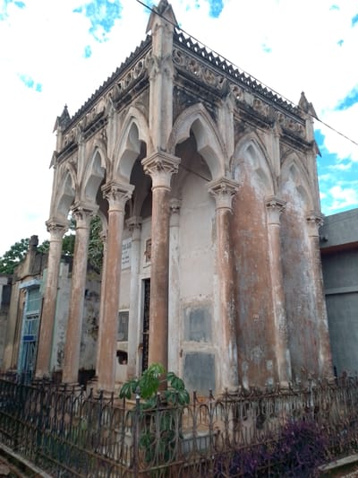 The image shows a weathered mausoleum in the Recoleta Cemetery of Asunción, Paraguay. The structure features a Gothic Revival architectural style, characterized by pointed arches, columns with Corinthian capitals, and decorative tracery along the roofline. The mausoleum is constructed of light-colored stone, with significant staining and weathering evident on the surfaces. A wrought-iron fence surrounds the structure, with plants growing both inside and outside the fence. A neighboring structure with a blue window is visible on the left, while a modern building stands on the right. The sky is visible in the background, with a blue sky scattered with white clouds.